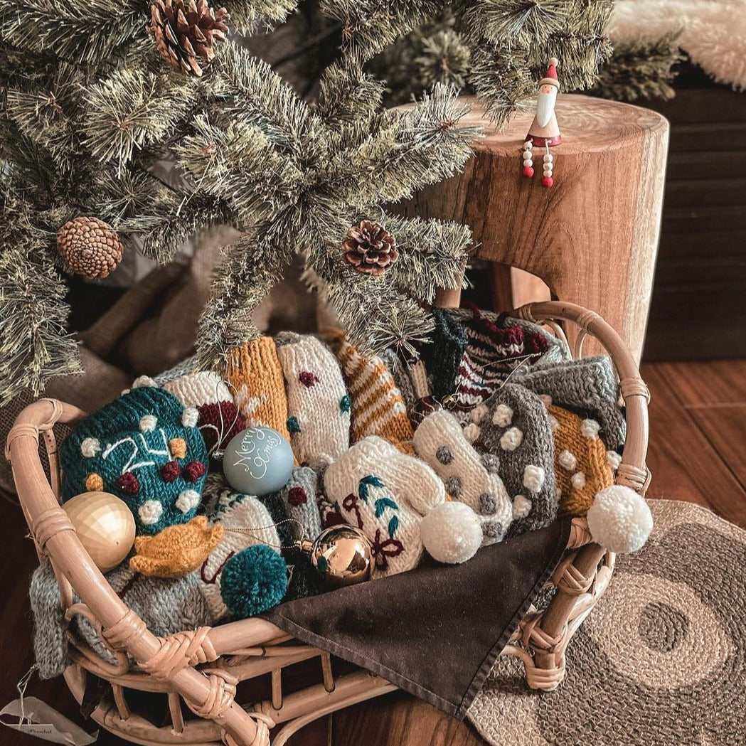 Handcrafted Christmas stockings, hats, and mittens arranged in a basket beneath a Christmas tree.