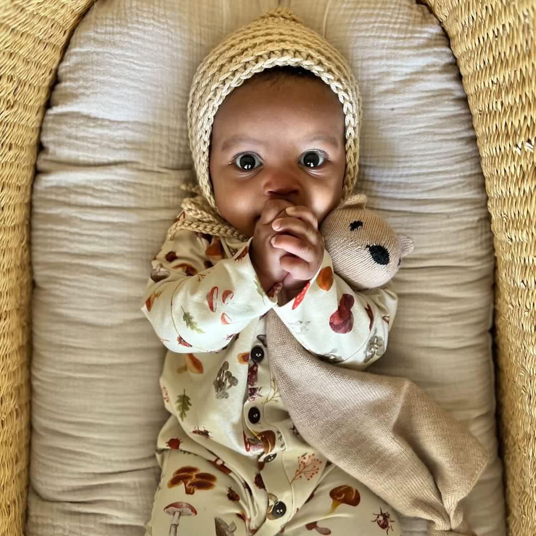 A baby dressed in the mushroom-print romper and a knitted bonnet cuddles a soft toy while lying in a basket.