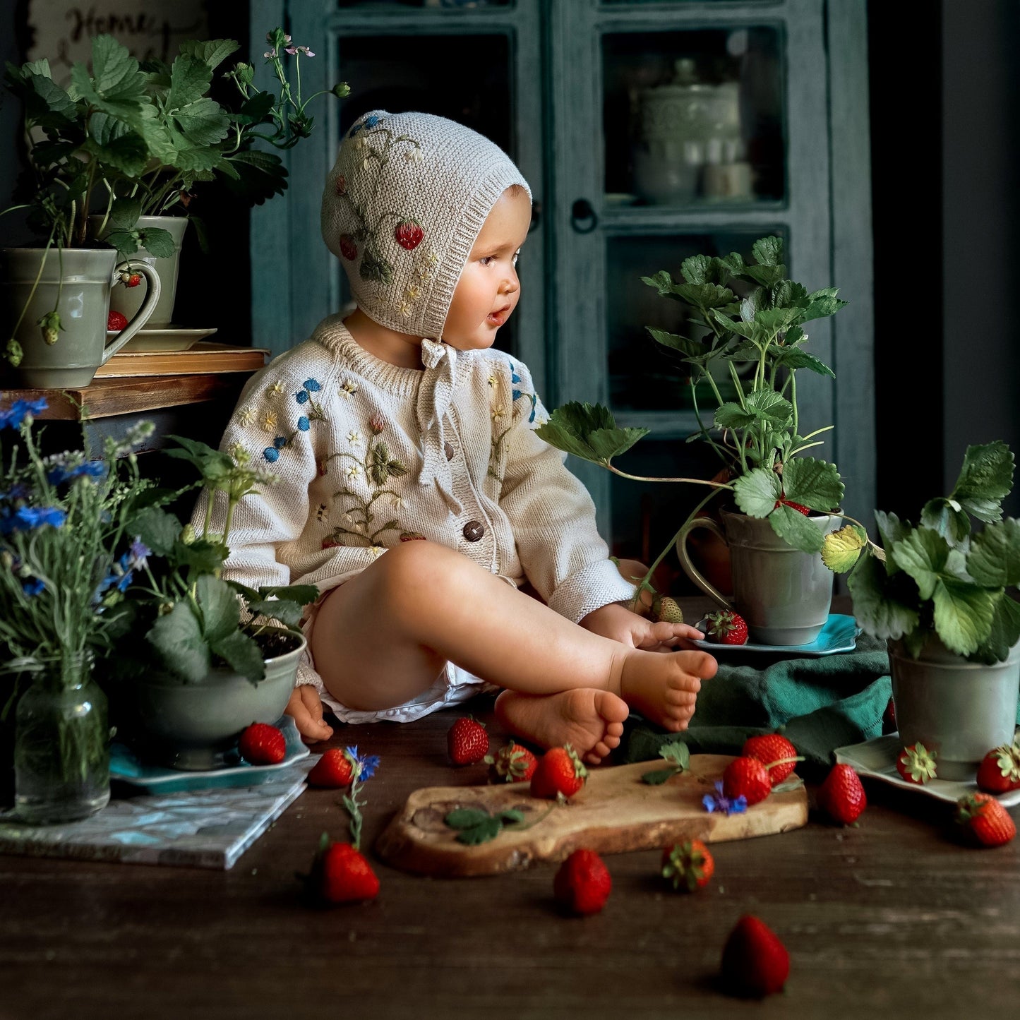 Child wearing a knitted bonnet with embroidered strawberries surrounded by plants and strawberries
