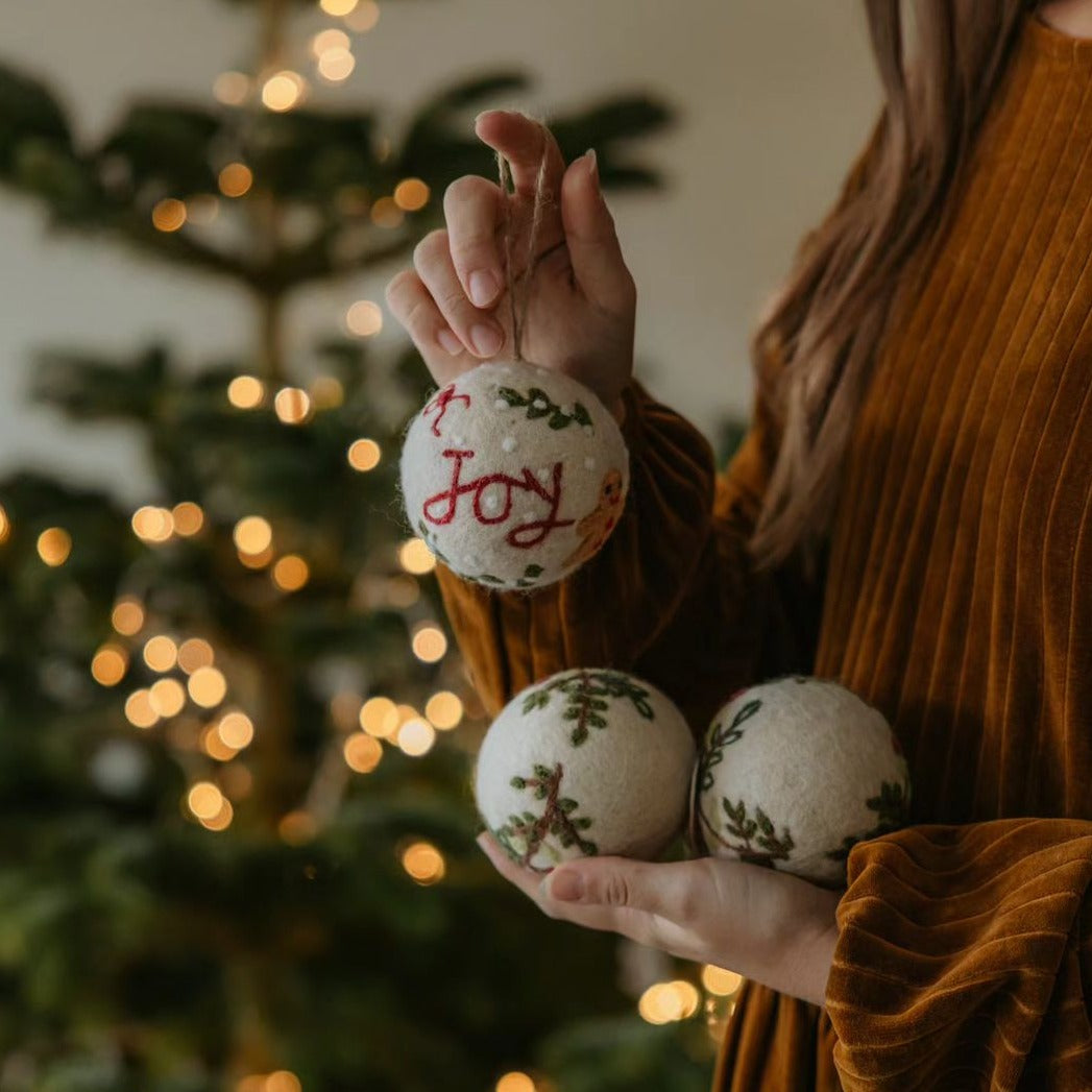 Multiple bauble ornaments with embroidered wreath designs, with a dimly lit Christmas tree in the background