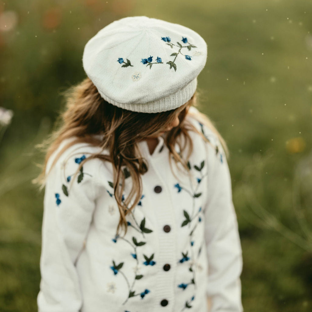 Person wearing a white embroidered beret and cardigan in a natural setting