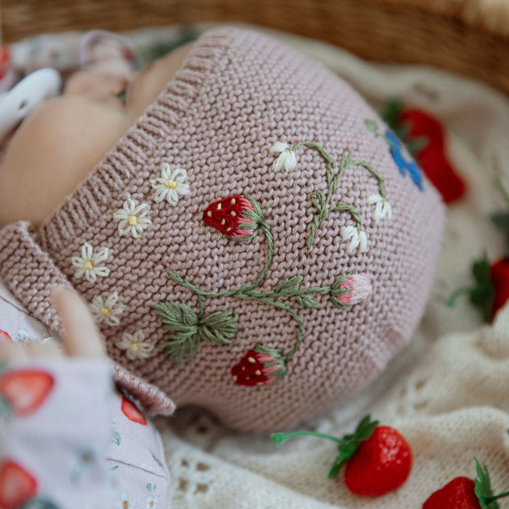 Pink knitted bonnet with floral embroidery placed beside purple and white flowers
