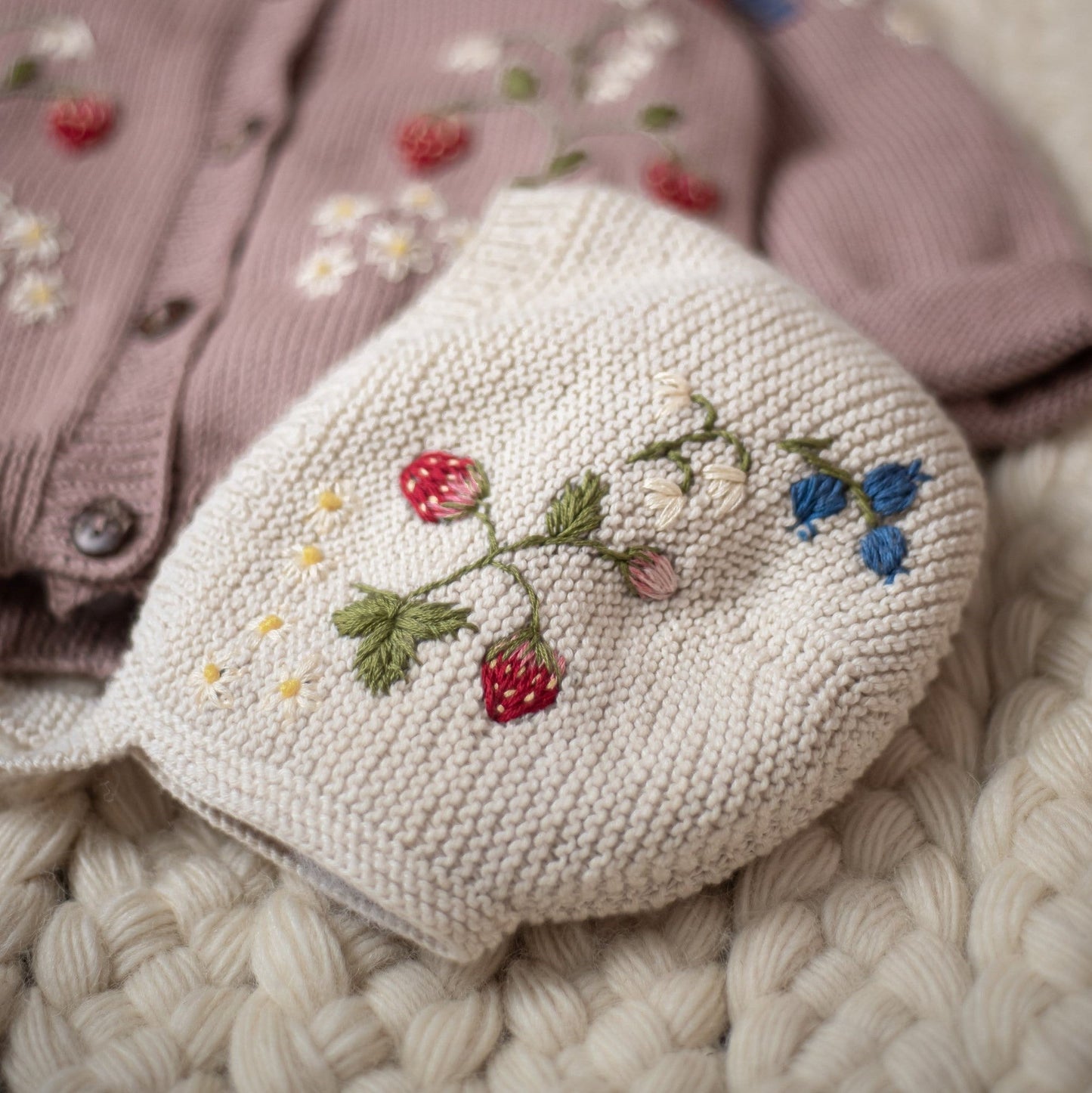 Close up of white knitted bonnet with embroidered strawberries and blue flowers beside a cardigan
