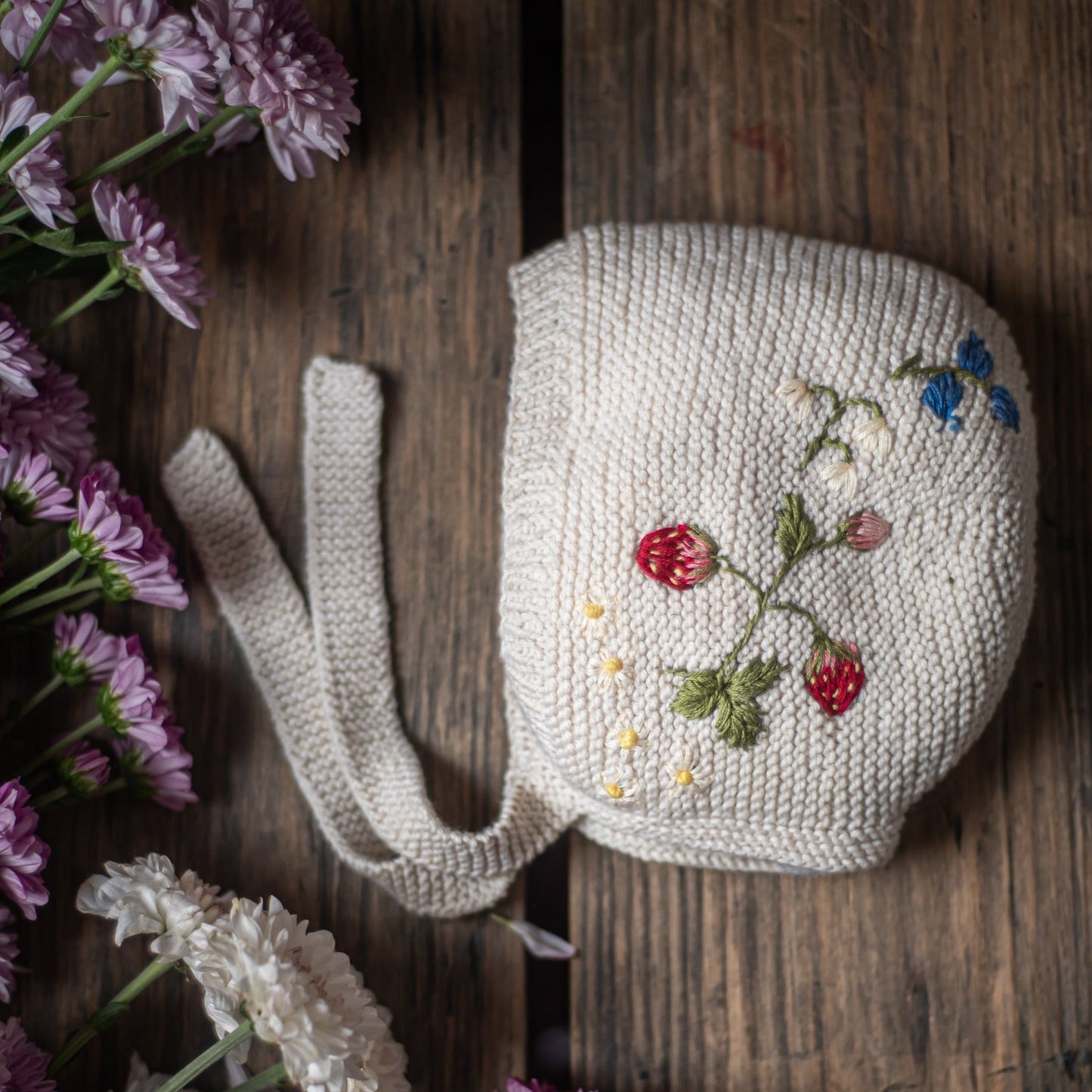 White knitted bonnet with embroidered strawberries and flowers on wooden table with blossoms