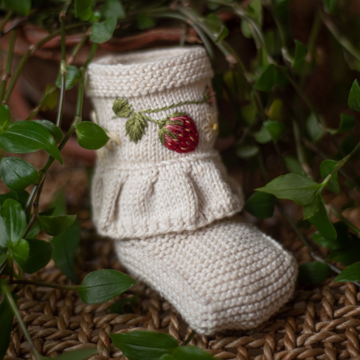 A single embroidered baby bootie resting among green leaves, emphasizing its natural elegance and handmade artistry.