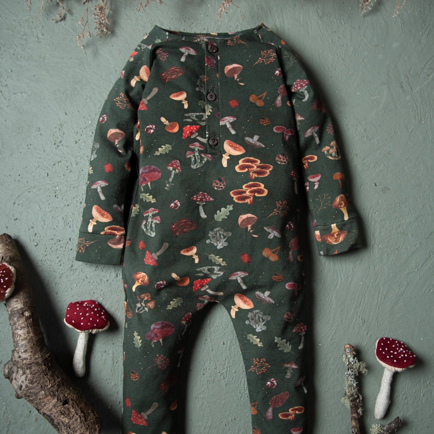 A dark green mushroom-print baby romper displayed against a textured green wall with toy mushrooms beside it.