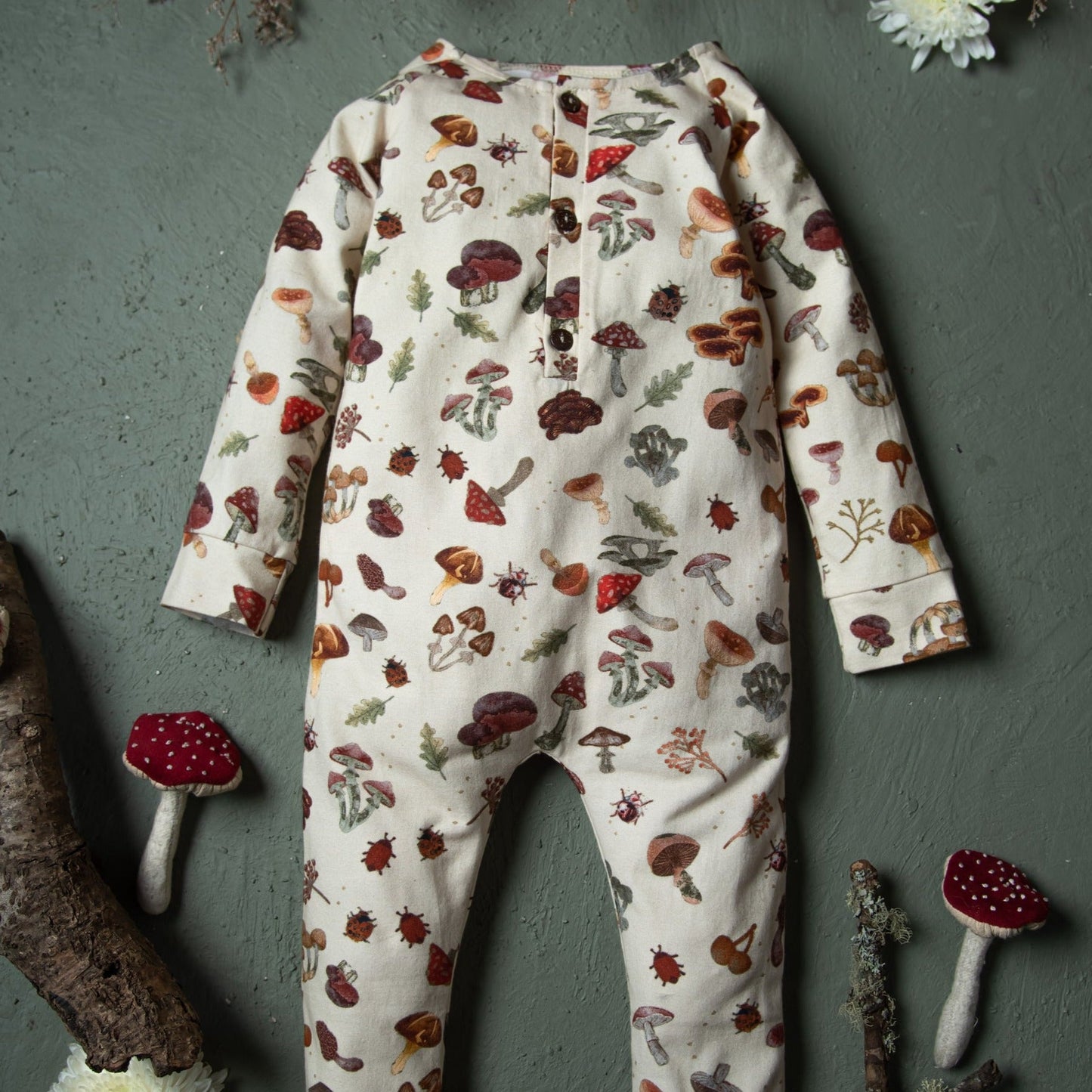 The cream mushroom-print romper is laid flat on a textured green surface surrounded by fabric mushrooms.
