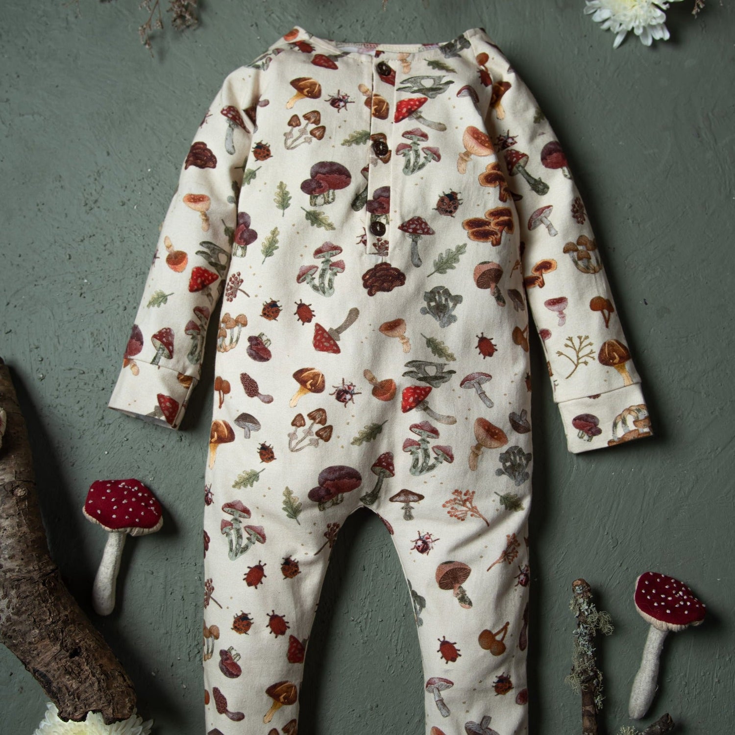 The cream mushroom-print romper is laid flat on a textured green surface surrounded by fabric mushrooms.
