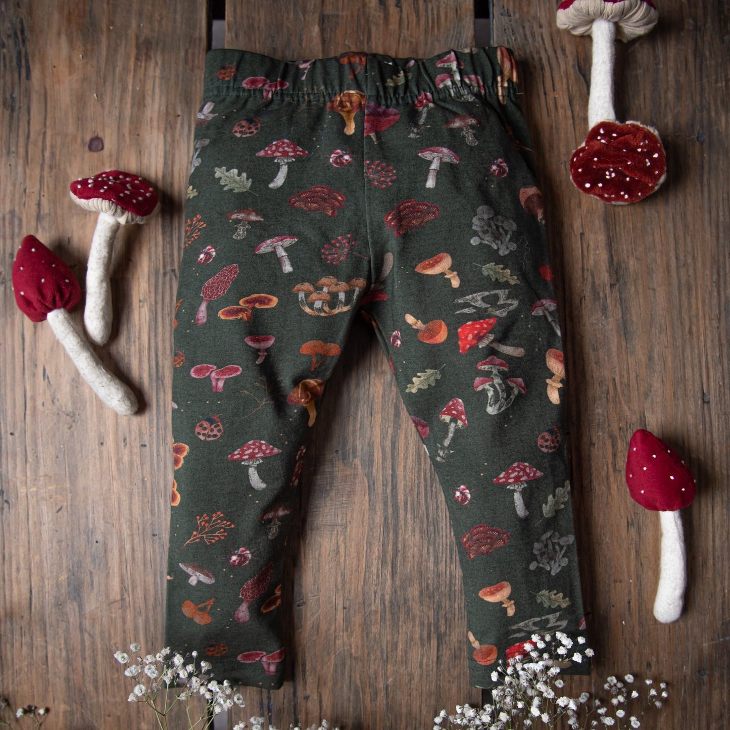 Mushroom-print leggings are styled flat on wood with toy mushrooms around.
