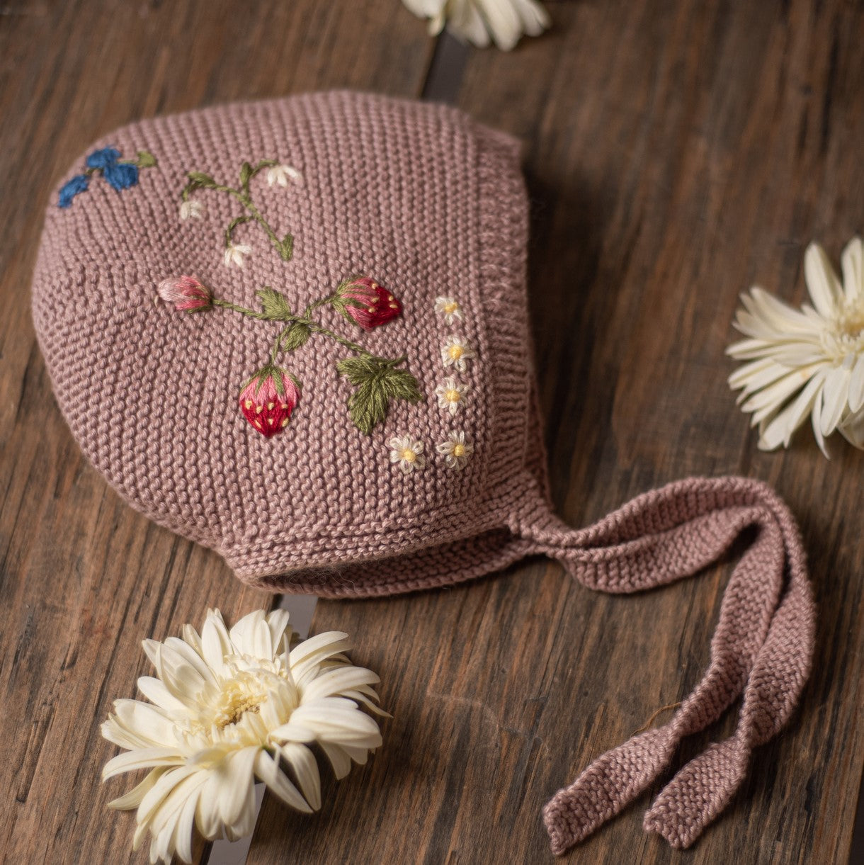 Close-up of pink knitted bonnet with embroidered strawberries and flowers on wooden background
