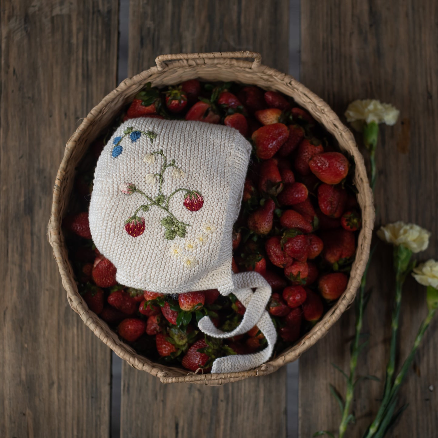 Knitted bonnet with strawberry embroidery placed in a basket of strawberries
