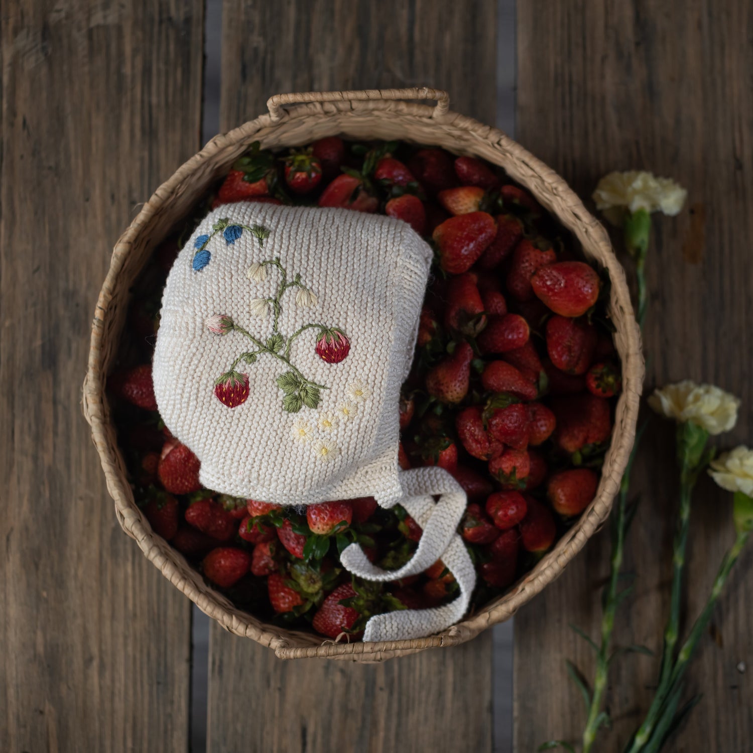 Knitted bonnet with strawberry embroidery placed in a basket of strawberries
