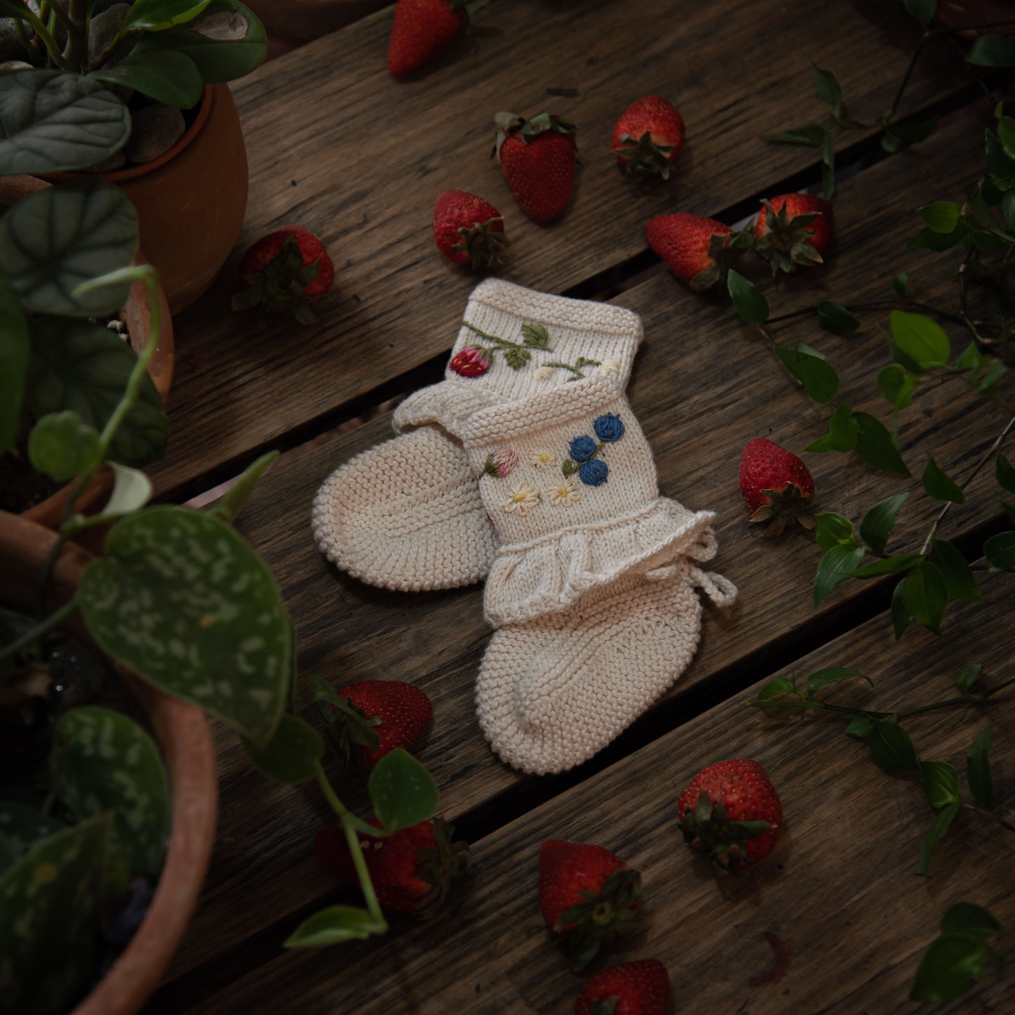 A rustic flatlay of embroidered baby booties surrounded by strawberries and greenery, capturing a cozy cottage charm.