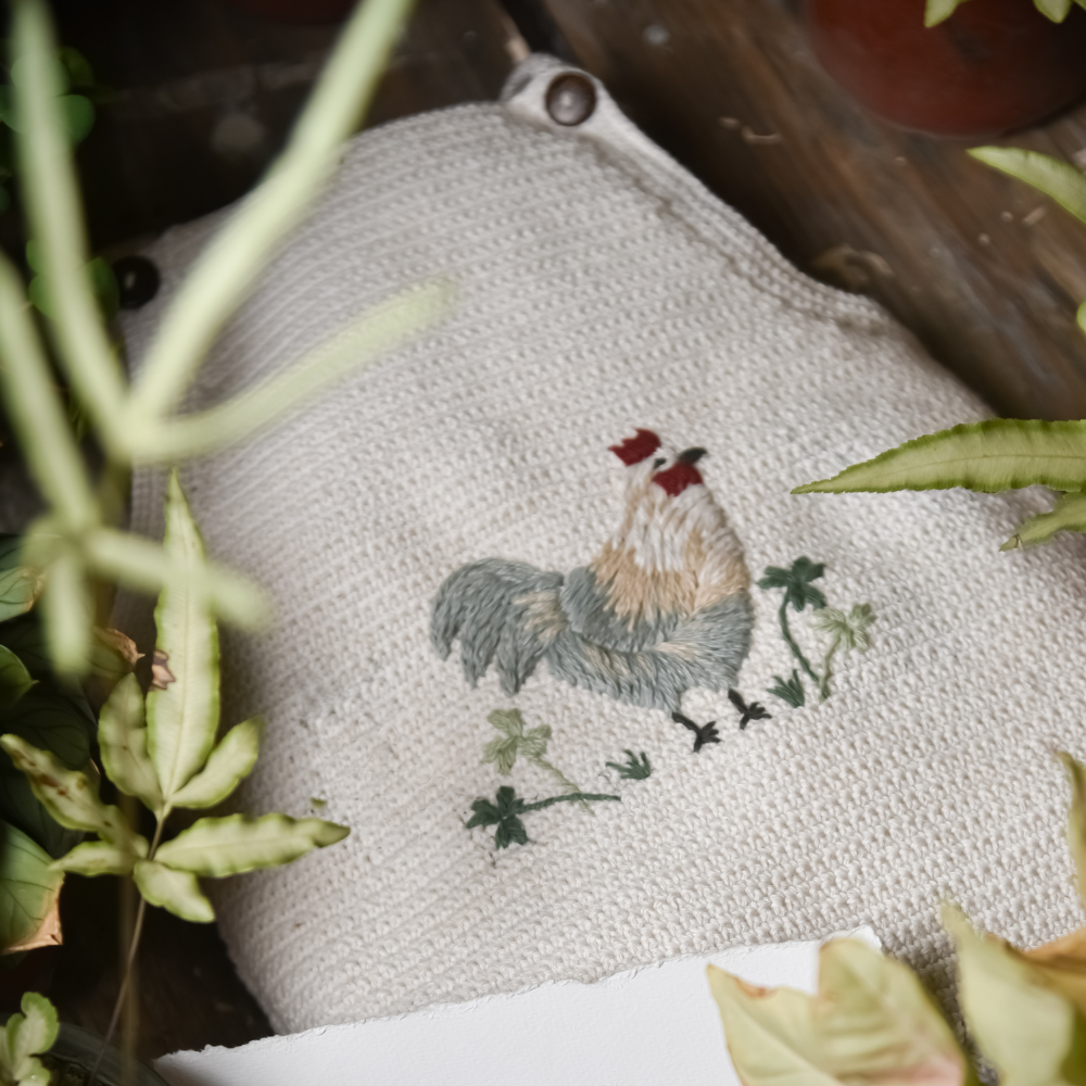 Close up of the Little Farm Romper - Malt with embroidered details, surrounded by greenery.