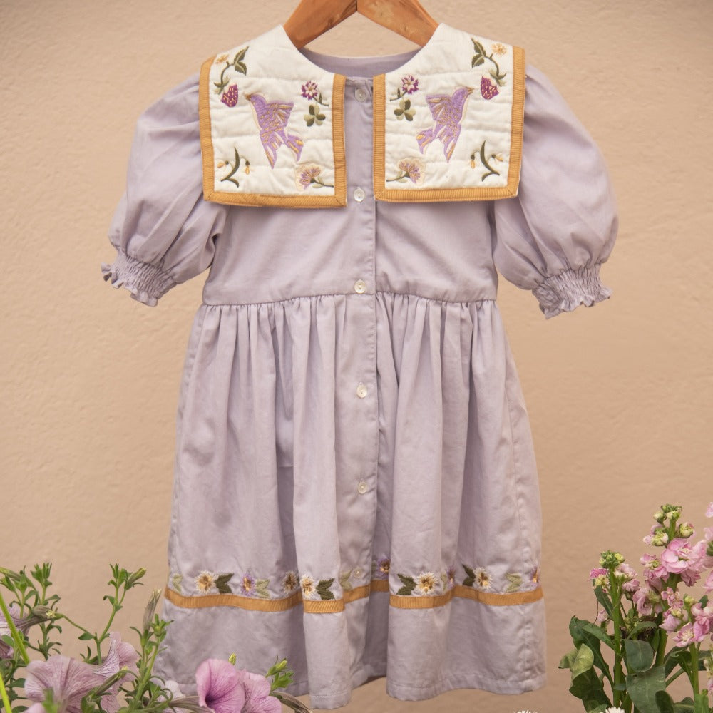 Birdie Bliss Dress - Lilac on a hanger, with flowers in the background.