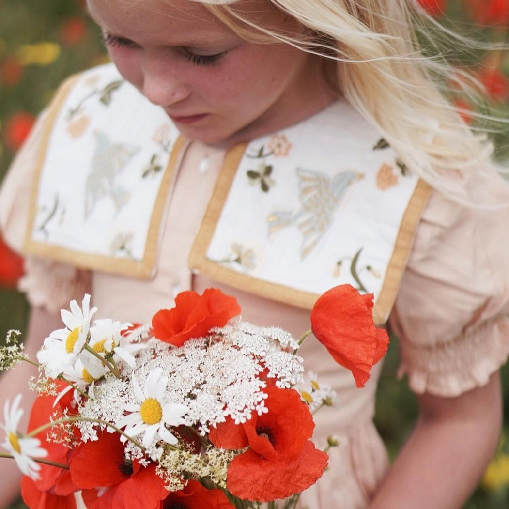 Girl wearing the Birdie Bliss dress in Apricot, surrounded by flowers.








