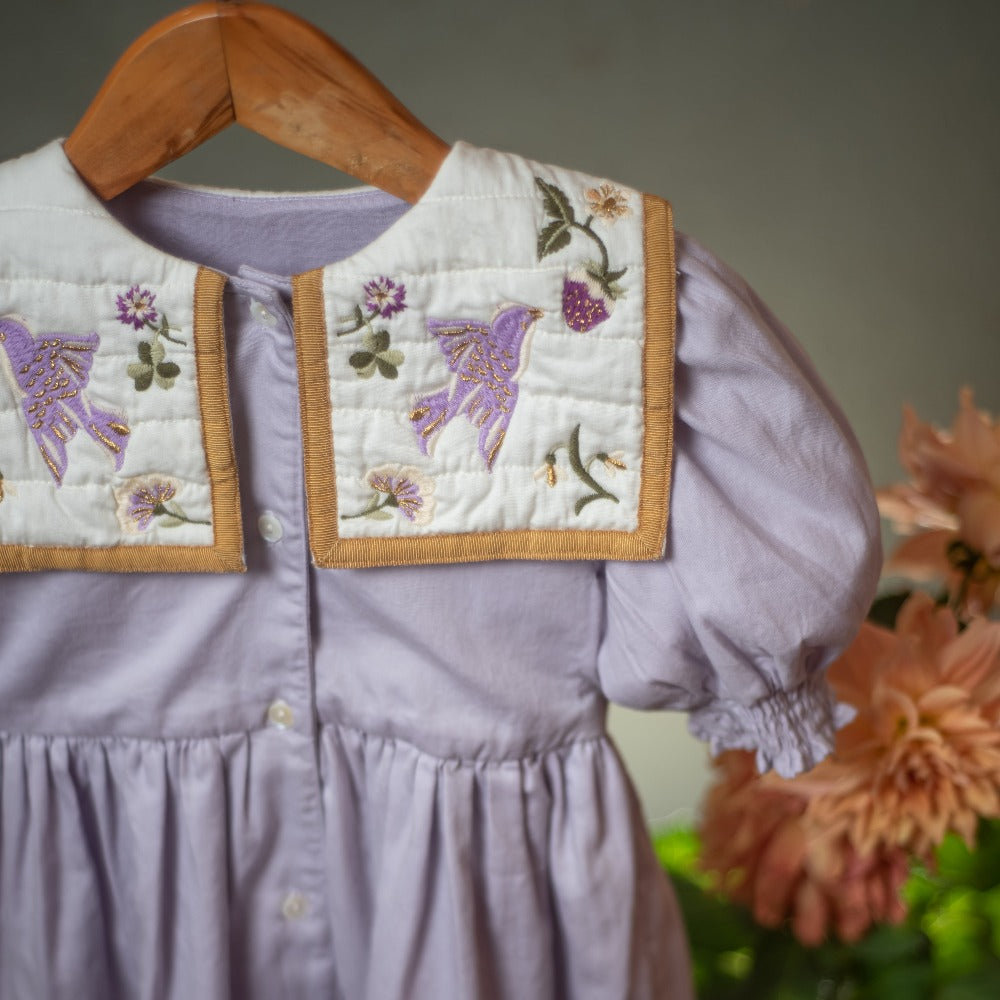 Part of the Birdie Bliss Dress - Lilac on a hanger, with faint flowers in the background.
