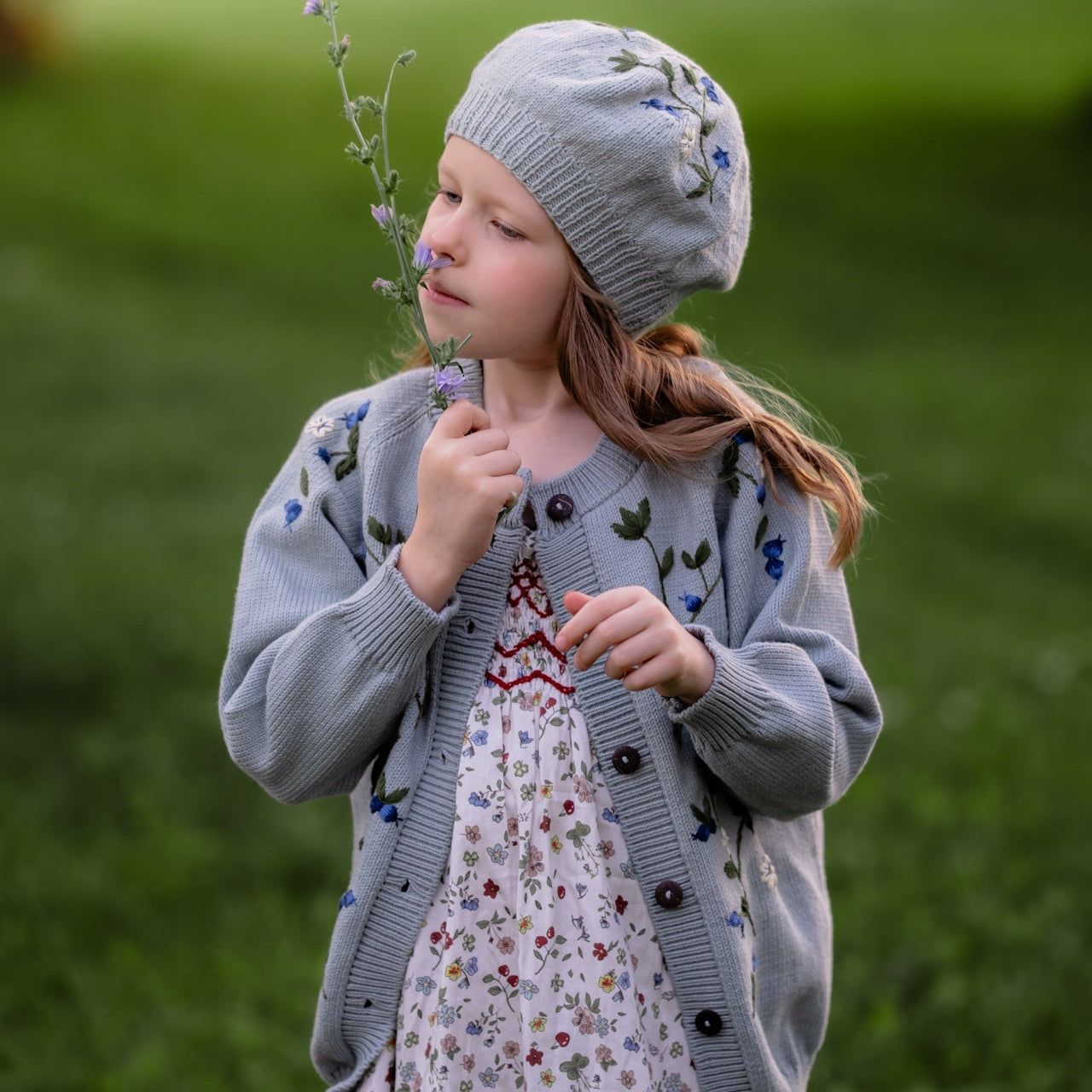 Young girl in a blue cardigan and floral dress holding a flower in a grassy field