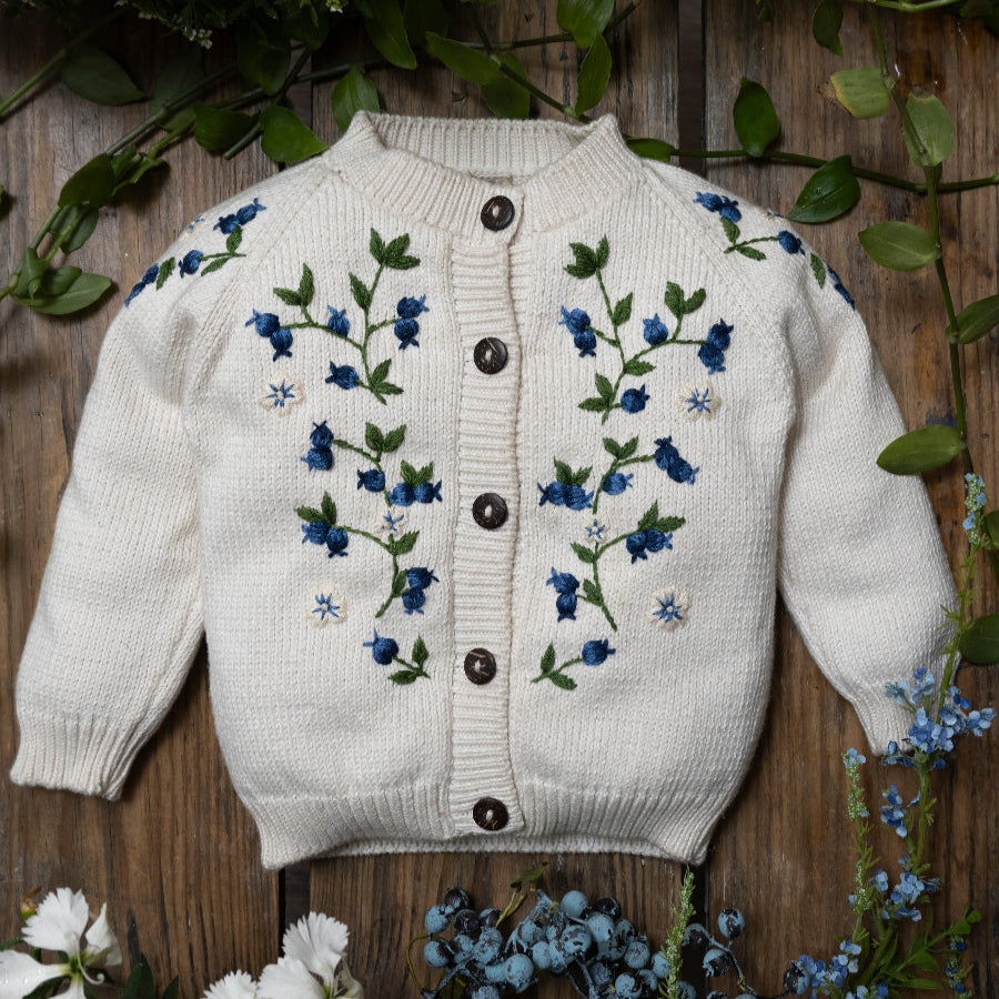 white knitted blueberries cardigan for kids displayed on a wooden surface surrounded with greenery 