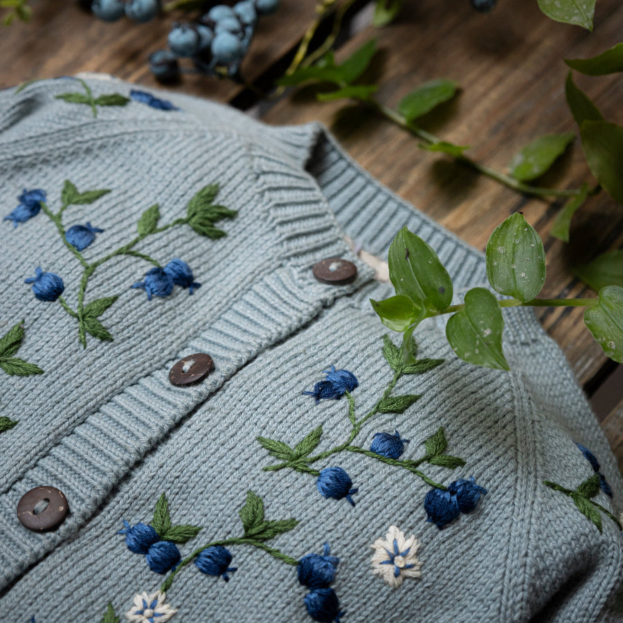 knitted and hand embroidered blueberries cardigan in Duck blue  displayed on a wooden surface