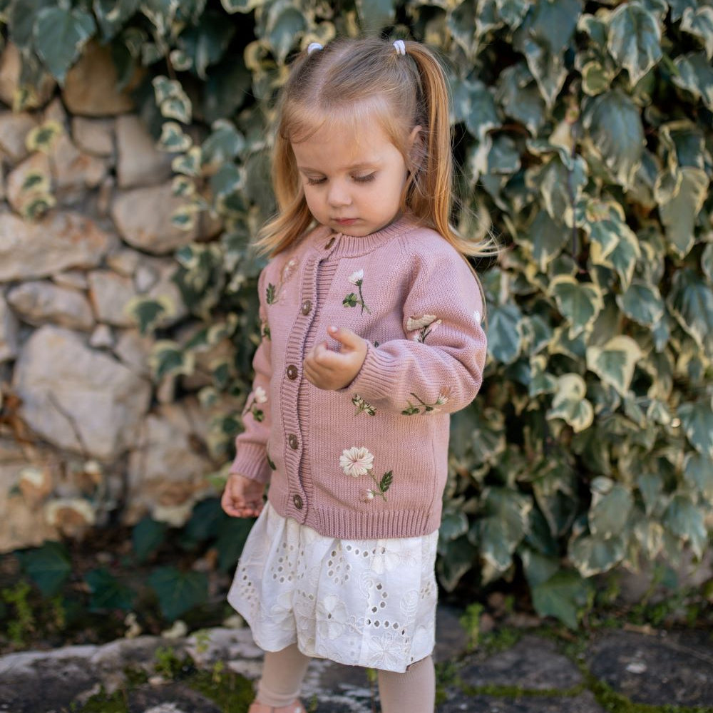 Young girl in a pink floral cardigan standing in front of a stone wall with greenery.