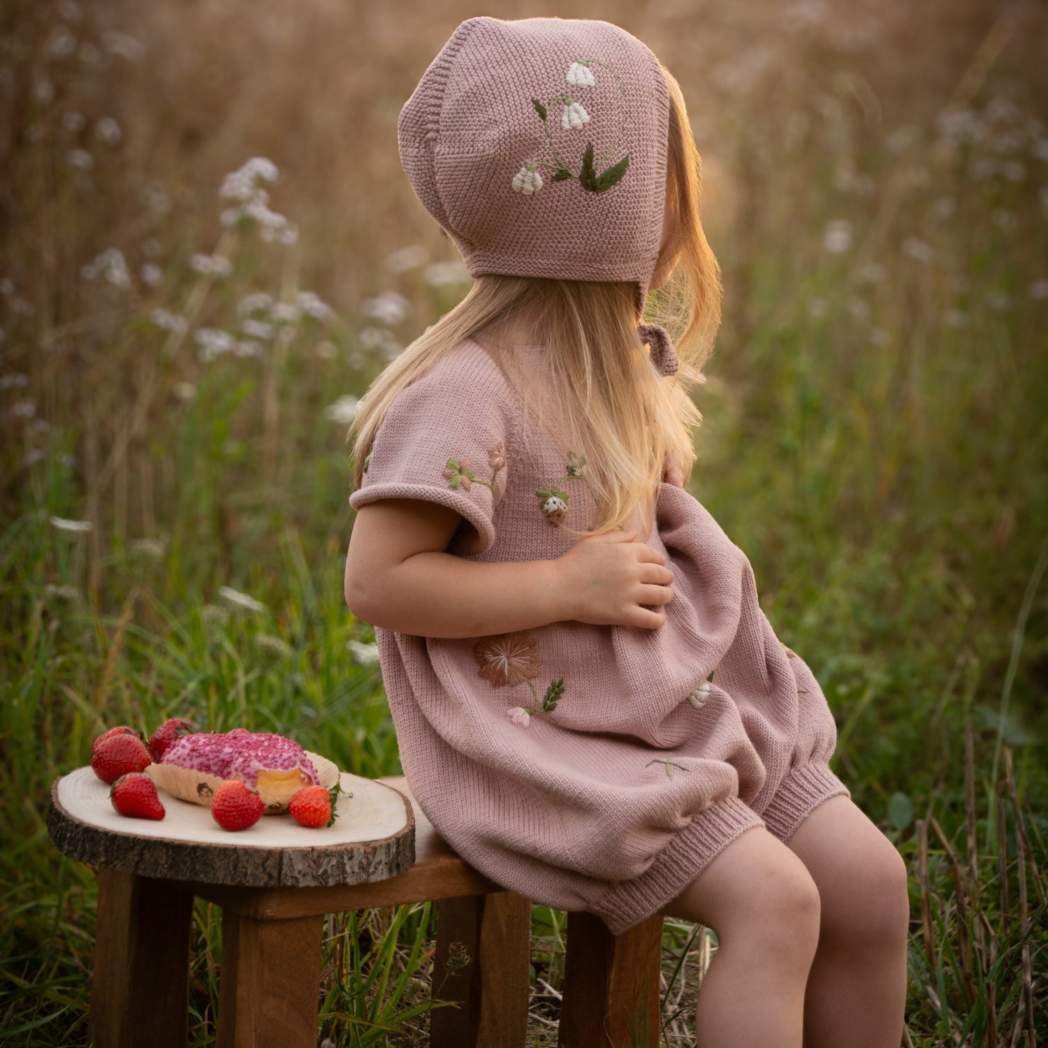 Child in a pink outfit sitting on a wooden stool in a field with a sunset.