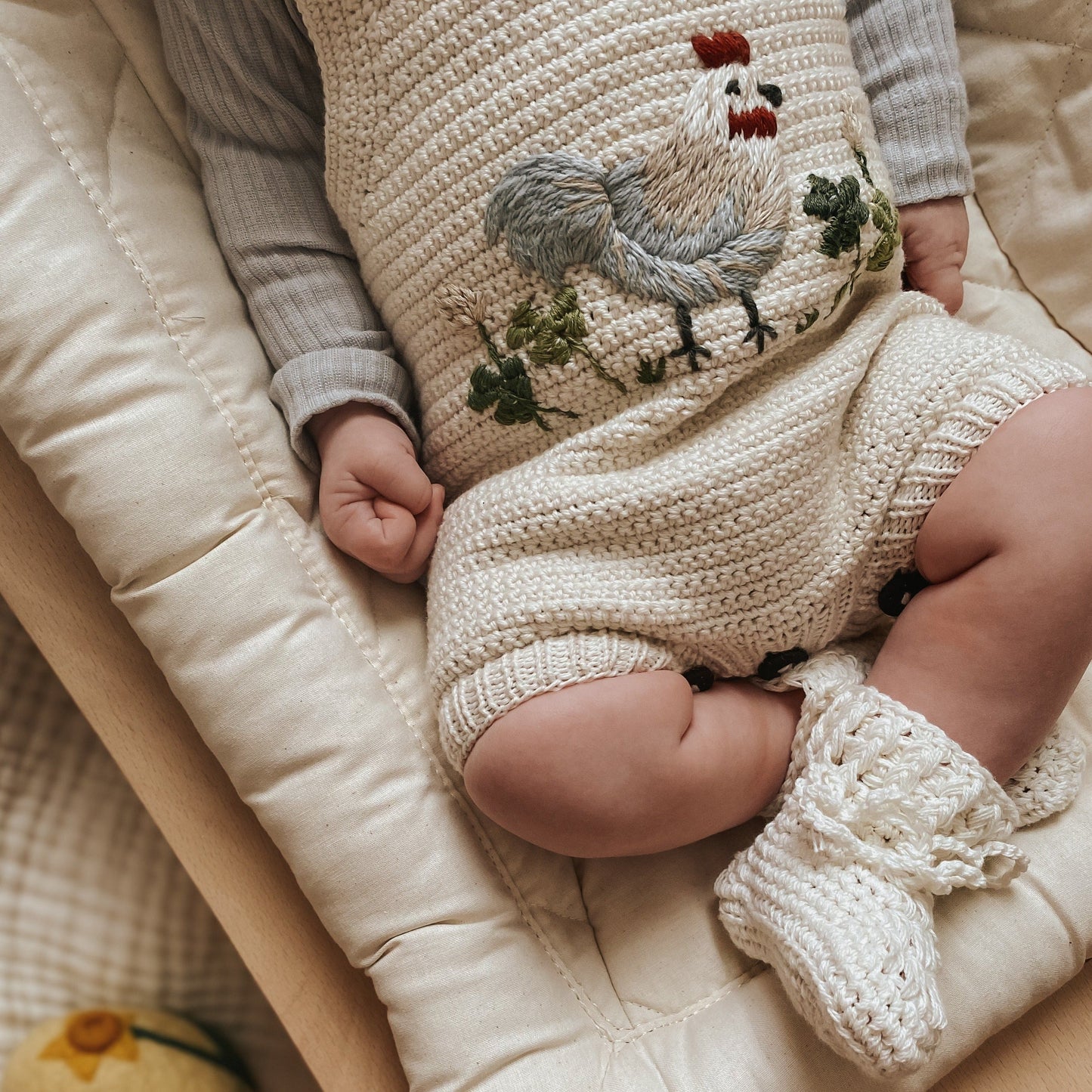 Baby wearing cream knitted romper with embroidered rooster and matching crochet booties






