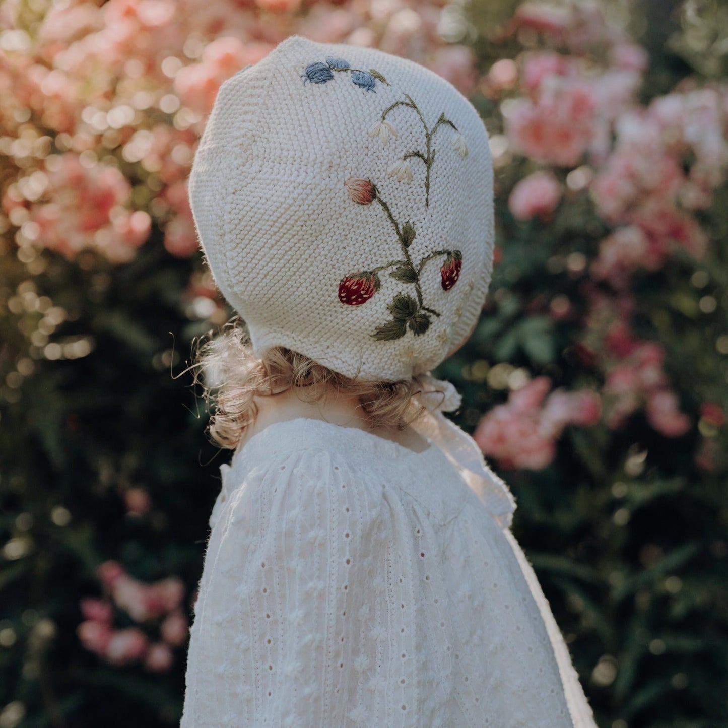 Girl wearing a white knitted bonnet with embroidered strawberries and flowers in a garden