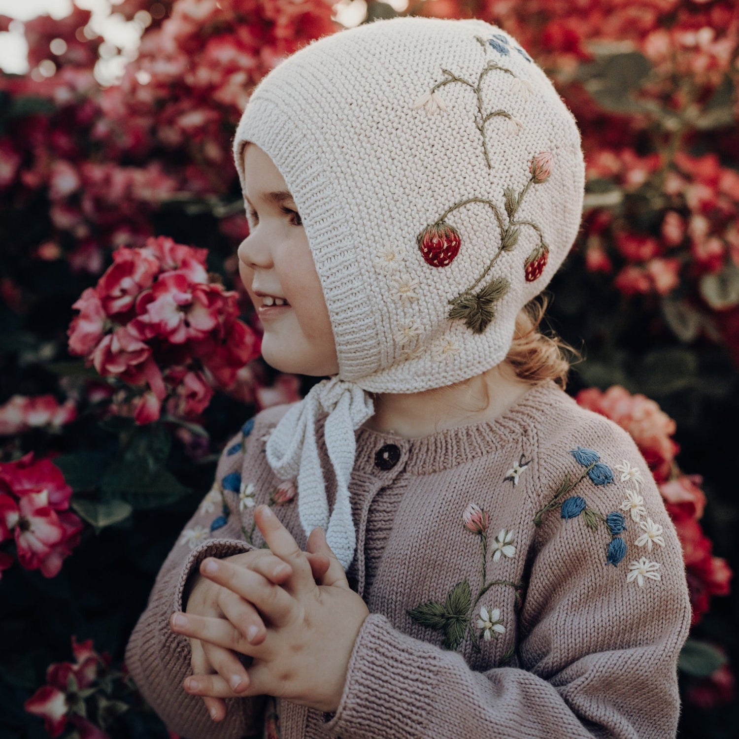Girl in white bonnet with embroidered strawberries standing by pink flowers
