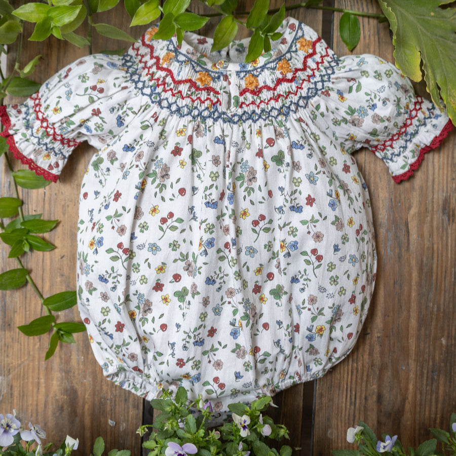 White baby romper with colorful floral print and red smocked embroidery on a wooden background