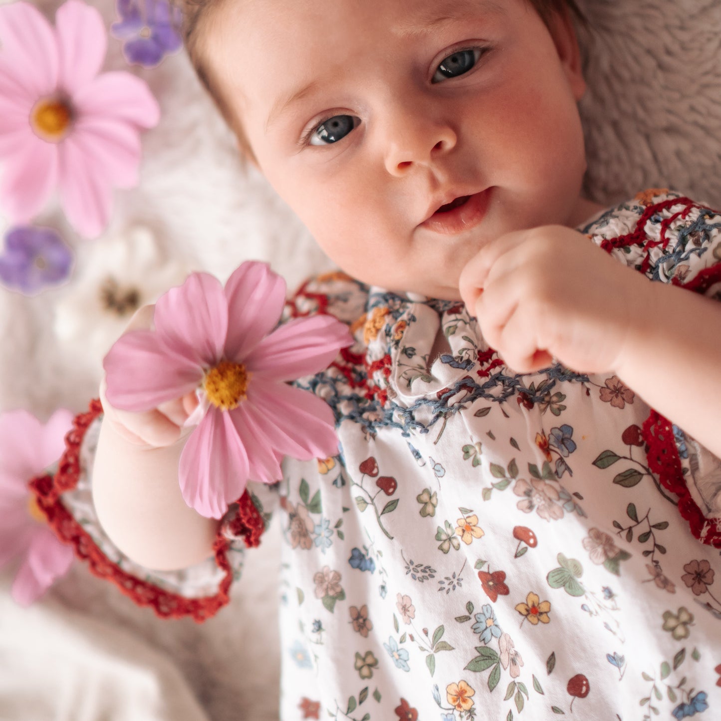 Baby lying on a soft surface with flowers around, wearing a floral dress.