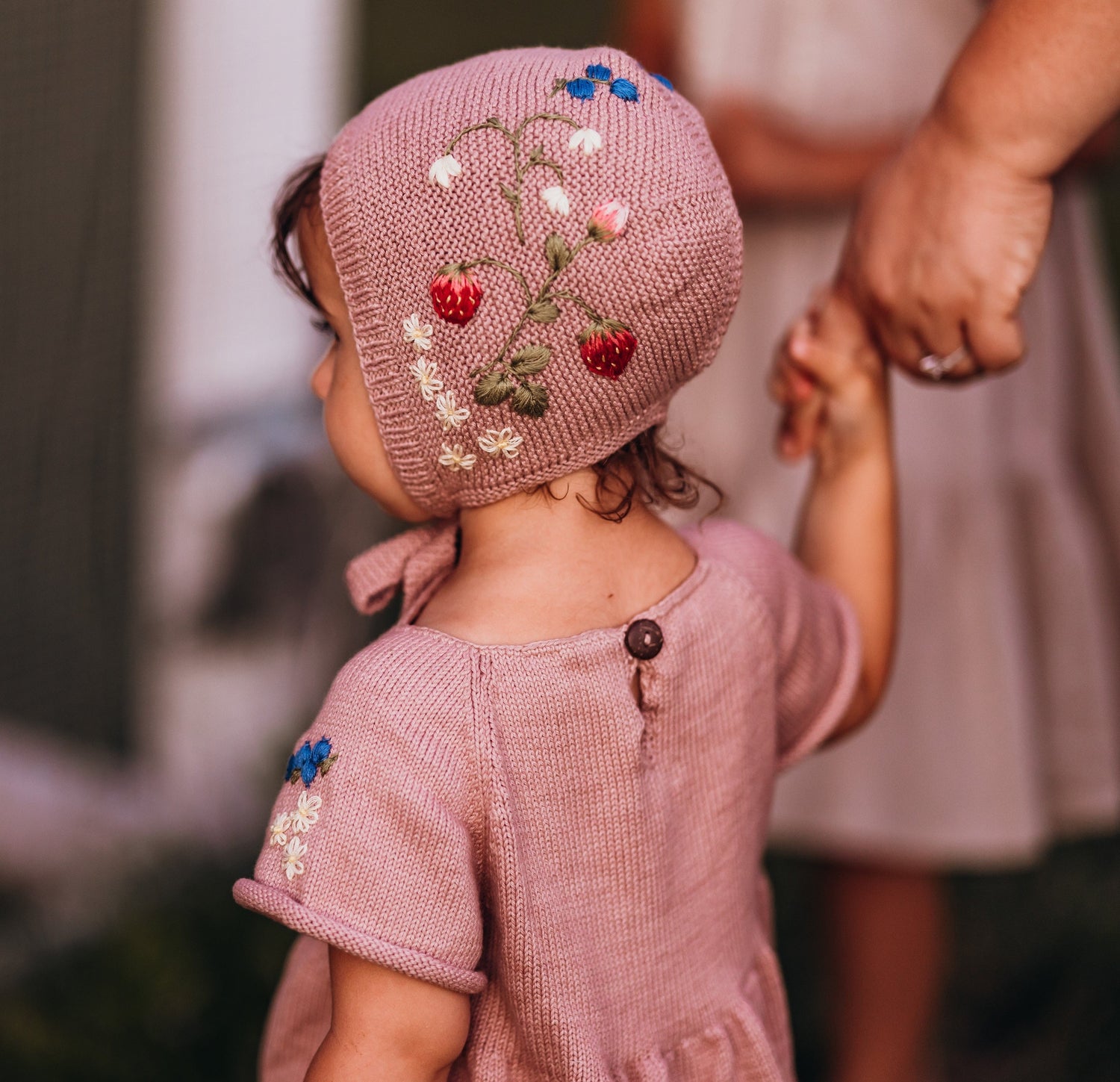 Pink knitted bonnet with floral embroidery placed on wooden surface with flowers
