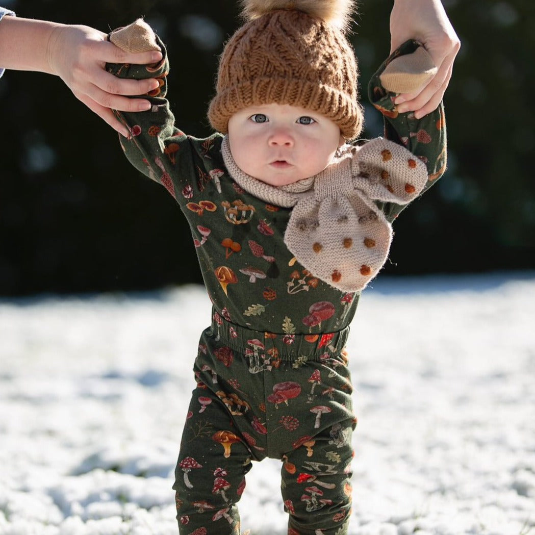 A baby is standing in the snow wearing the dark green mushroom set with winter accessories.