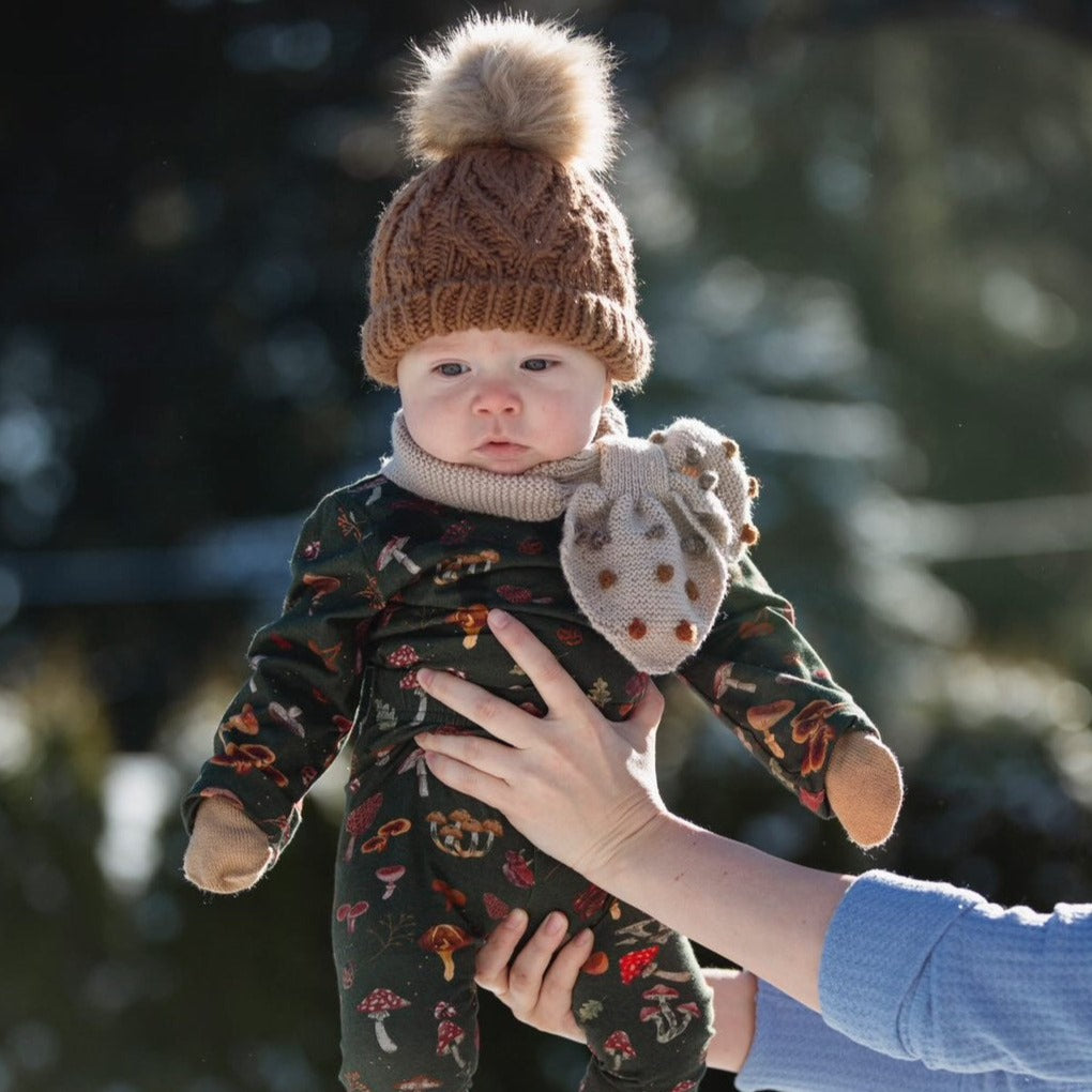 A baby outdoors wearing the mushroom-print outfit, a knit hat with a pom-pom, mittens, and a scarf.