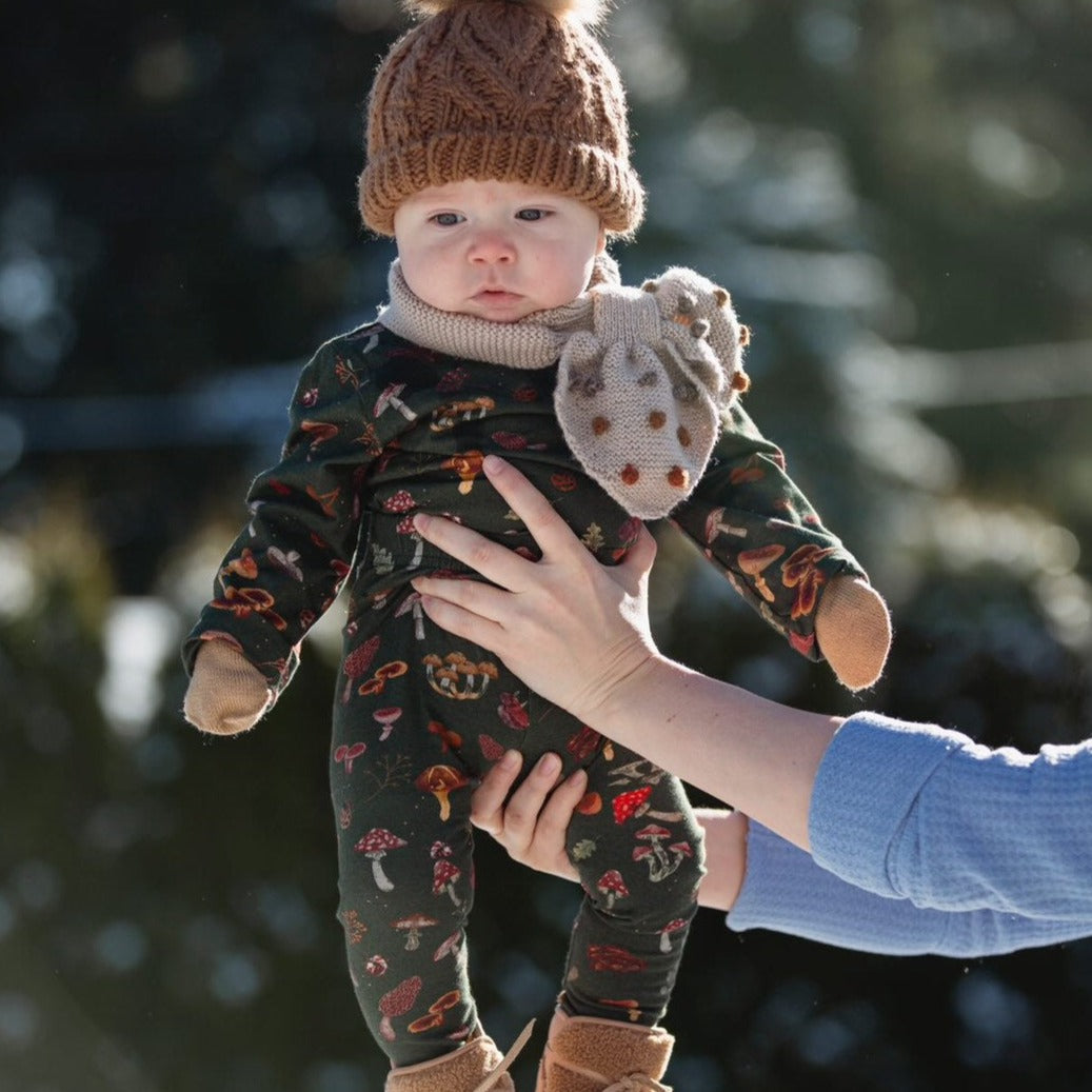 A baby is being held up outside, dressed in the dark green mushroom set with a knit hat and scarf.

