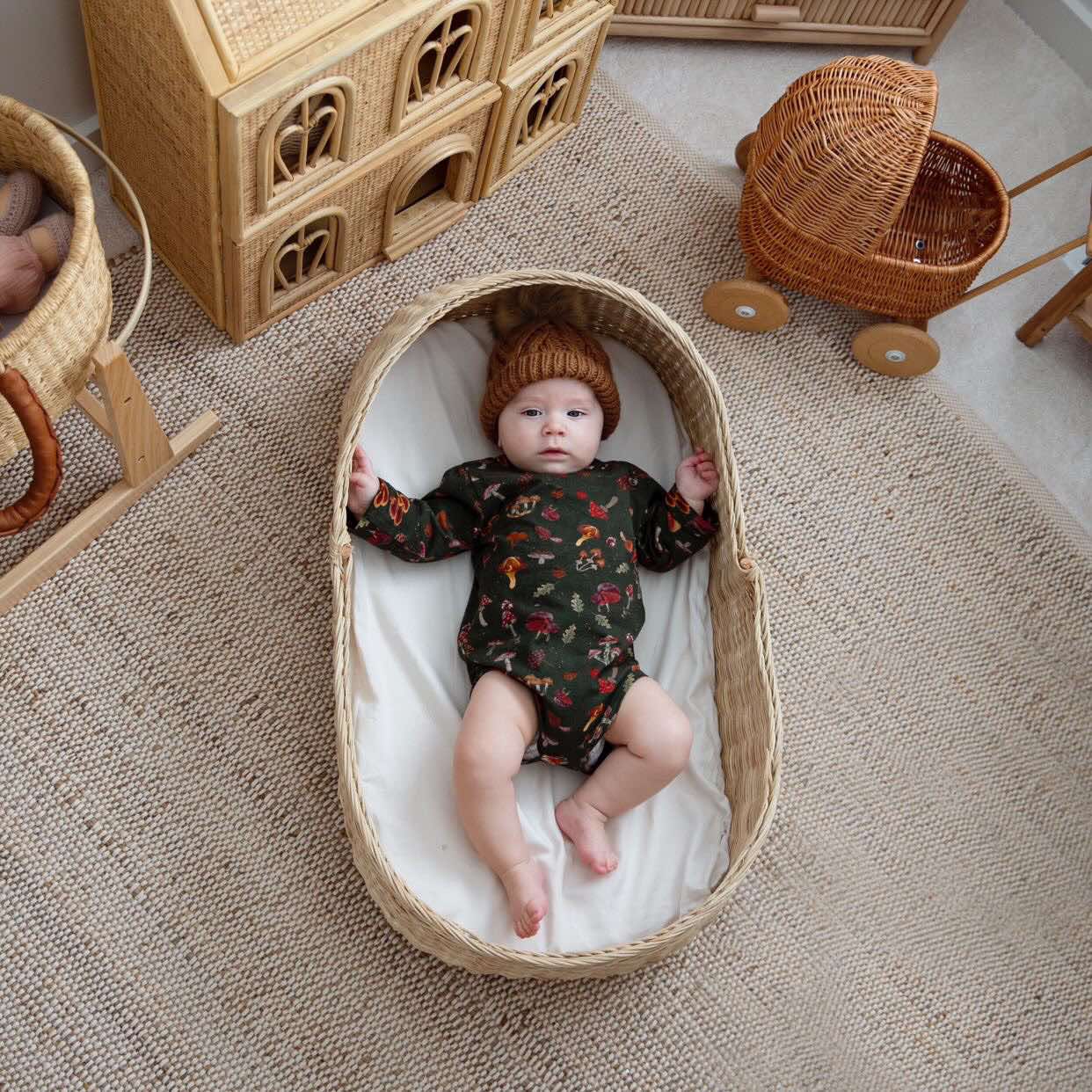 A baby in a wicker basket wearing the mushroom-print bodysuit and an amber knit hat.
