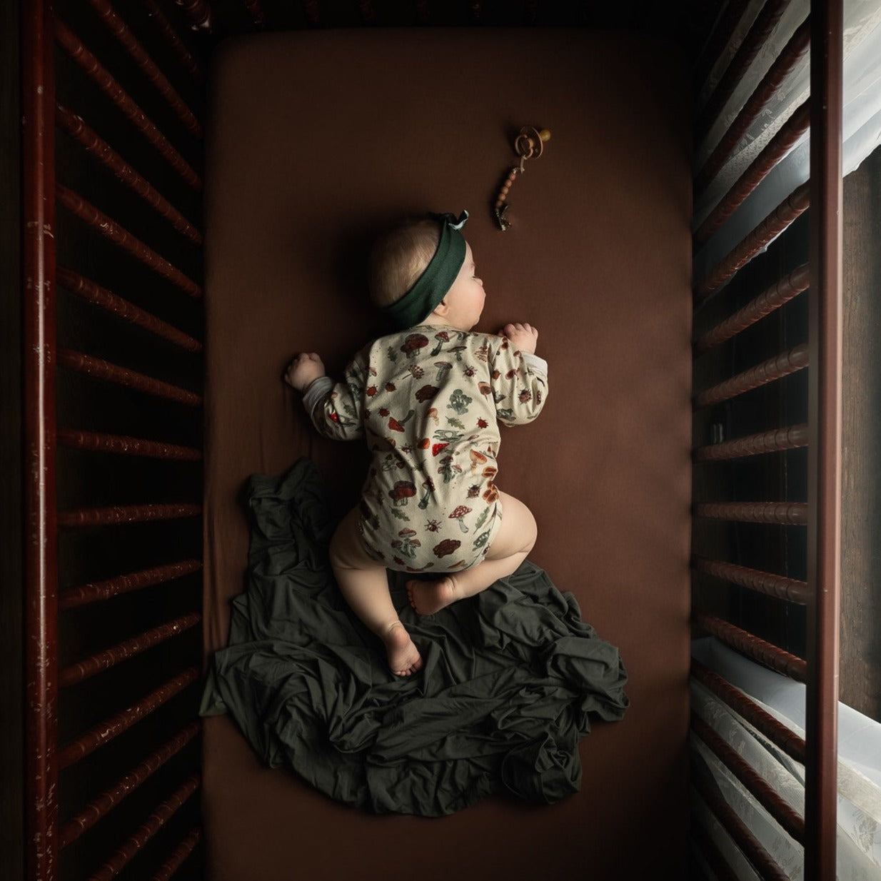 A baby sleeping on their stomach in a crib, dressed in the mushroom-print bodysuit with a green headband.