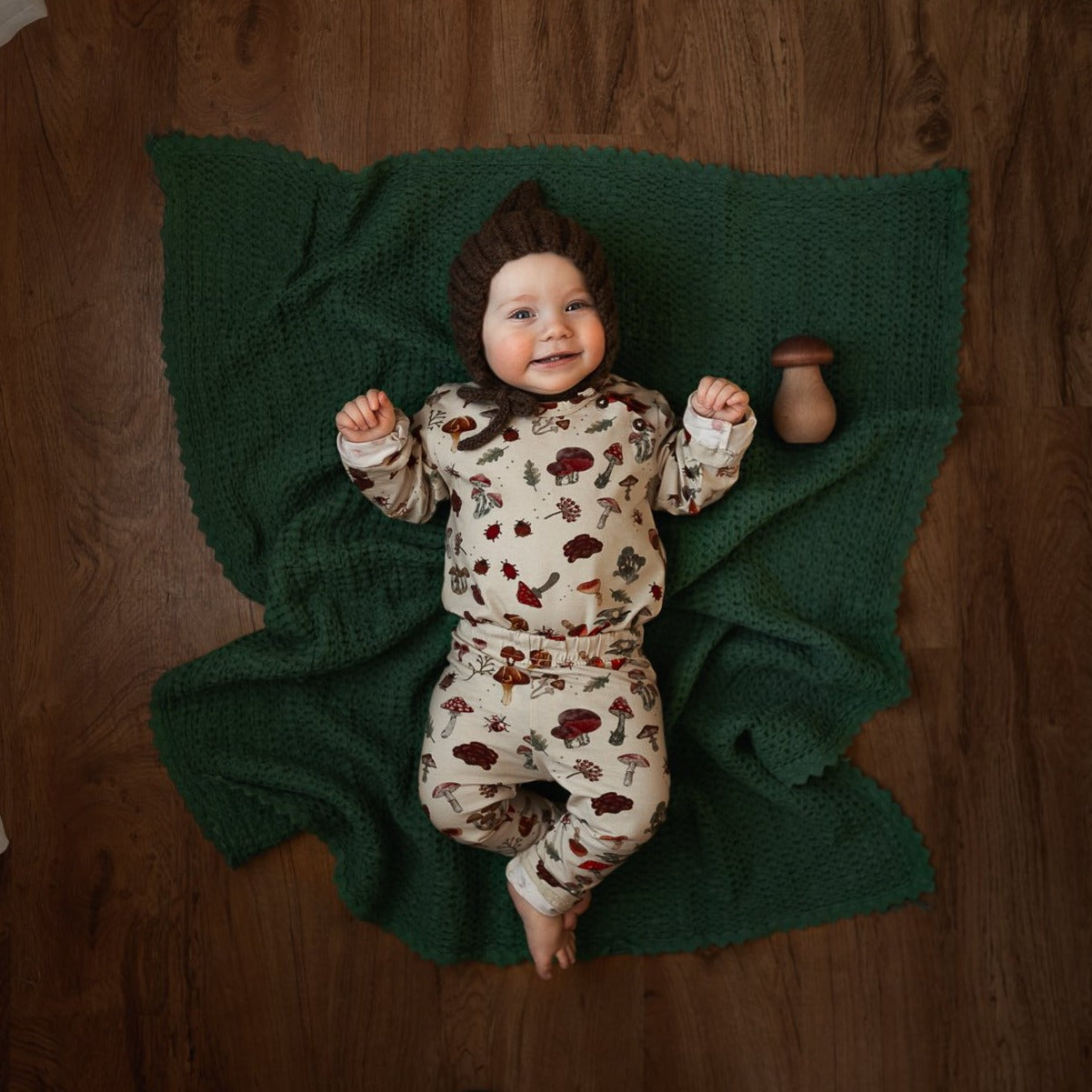 A smiling baby lies on a green blanket wearing a matching mushroom-print outfit and knitted bonnet.