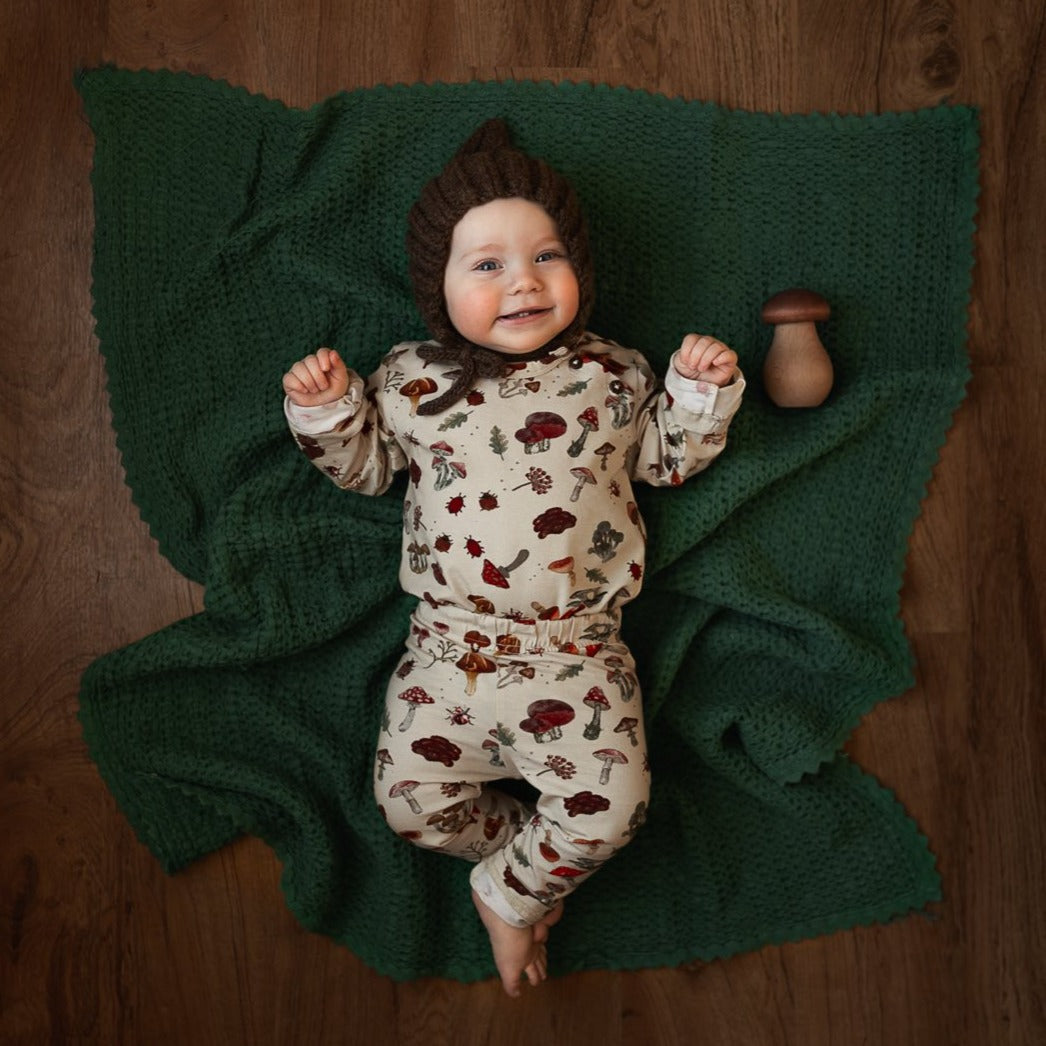 A baby smiling and lying on a green knit blanket while wearing a full outfit in the mushroom-print fabric.