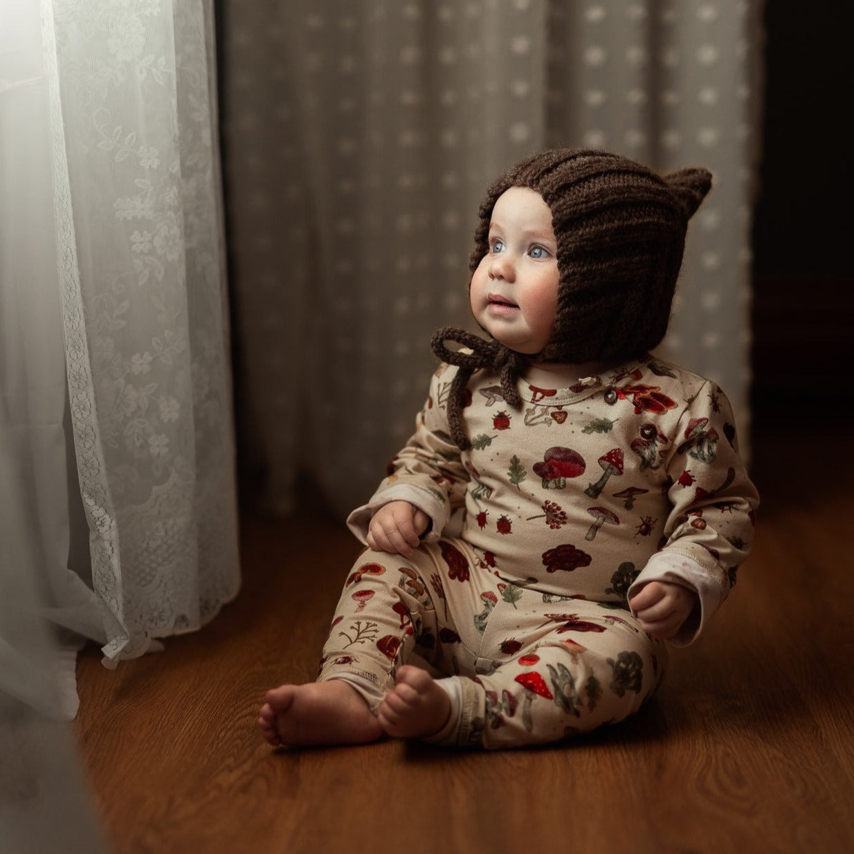 Baby in a cozy mushroom-print set sits by a window wearing a brown knitted bonnet.
