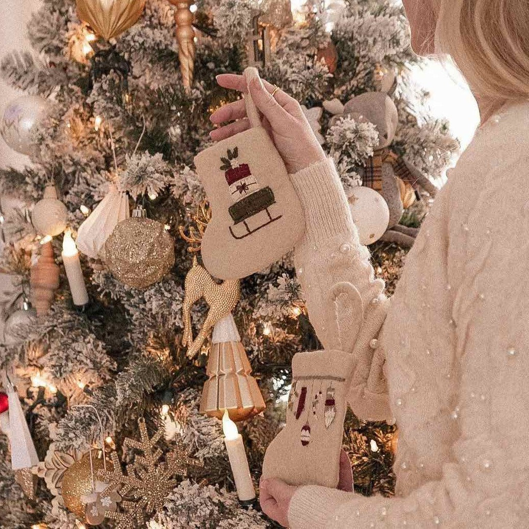 Multiple Mini Stocking Sleigh ornaments held in hands, positioned beside a Christmas tree adorned with various decorations.